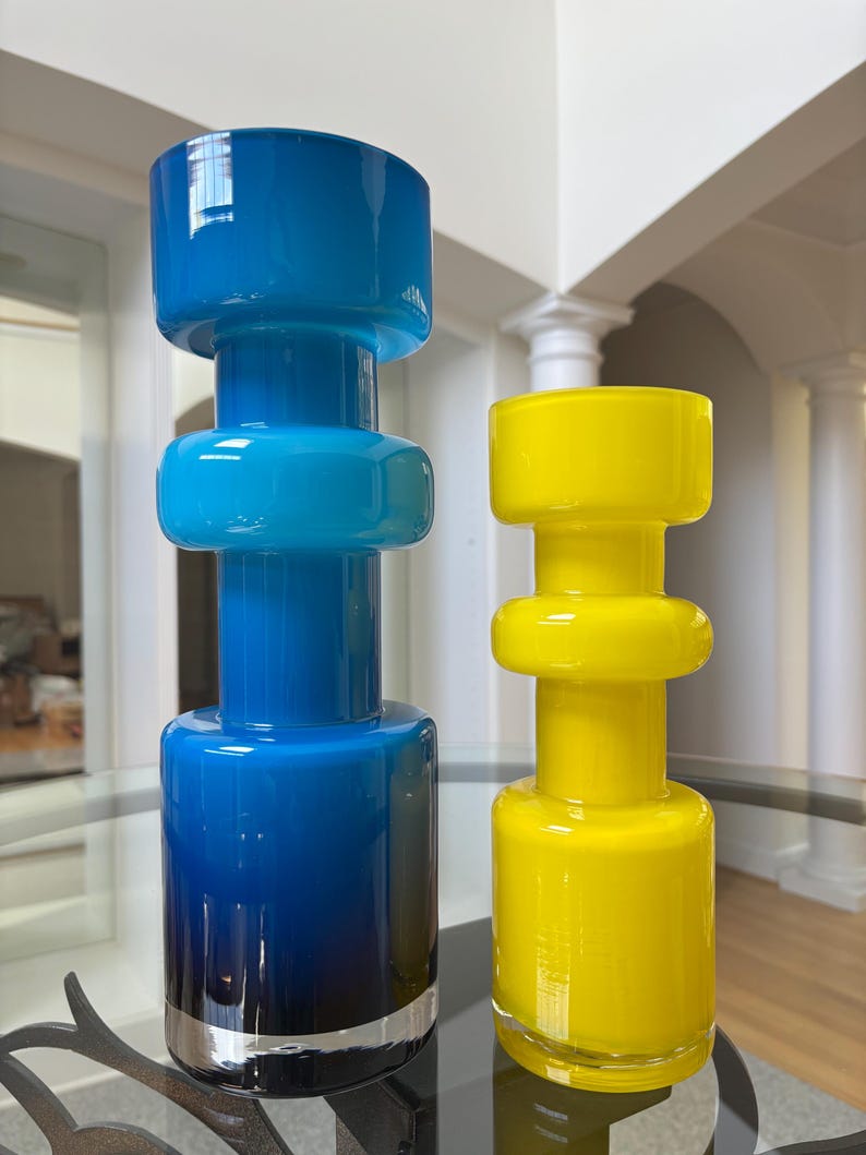 Peut inclure: Deux vases d&eacute;coratifs en verre, l'un bleu avec une base argent&eacute;e, l'autre jaune. Les deux ont une base cylindrique, une section centrale arrondie et un large dessus plat. Ils sont pos&eacute;s sur une table en verre.