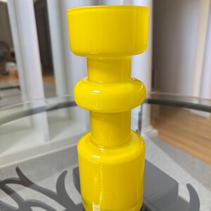 Peut inclure: Un vase en verre jaune vif avec un design g&eacute;om&eacute;trique unique. Le vase pr&eacute;sente une base cylindrique, une section centrale arrondie et un dessus large et rectangulaire. La finition brillante refl&egrave;te la lumi&egrave;re, soulignant la couleur vibrante et la texture lisse.