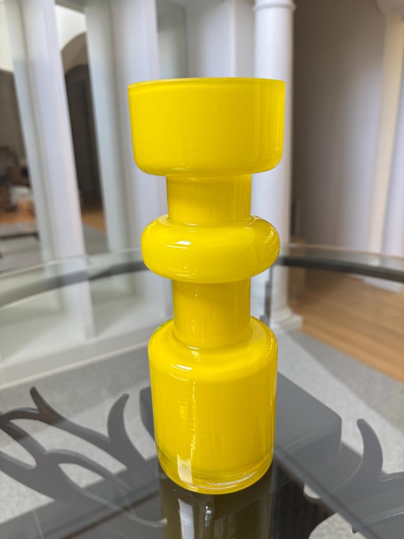 Peut inclure: Un vase en verre jaune vif avec un design g&eacute;om&eacute;trique unique. Le vase pr&eacute;sente une base cylindrique, une section centrale arrondie et un dessus large et rectangulaire. La finition brillante refl&egrave;te la lumi&egrave;re, soulignant la couleur vibrante et la texture lisse.