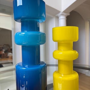Peut inclure: Deux vases d&eacute;coratifs en verre. L'un est un d&eacute;grad&eacute; de bleu, passant d'un indigo profond &agrave; la base &agrave; un bleu ciel vif au sommet. L'autre est jaune vif. Les deux ont une base cylindrique, une section centrale arrondie et un dessus large et plat.