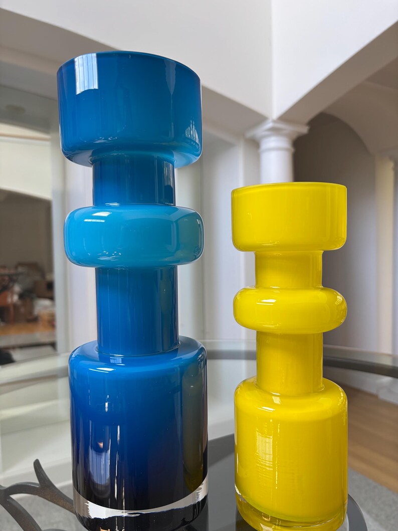 Peut inclure: Deux vases d&eacute;coratifs en verre. L'un est un d&eacute;grad&eacute; de bleu, passant d'un indigo profond &agrave; la base &agrave; un bleu ciel vif au sommet. L'autre est jaune vif. Les deux ont une base cylindrique, une section centrale arrondie et un dessus large et plat.