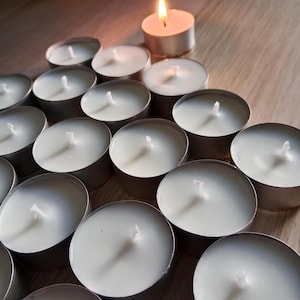 Puede incluir: Un grupo de velas de té blancas en recipientes plateados dispuestos en forma de pirámide sobre una superficie de madera. Una sola vela de té encendida se encuentra en la parte superior de la pirámide.