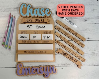 May include: A wooden first day of school sign with a light blue name cutout for Chase and a purple name cutout for Emersyn. The board features white dry-erase sections for grade, date, age, height, and learning goals, plus five interchangeable wooden strips with engraved prompts and a dry-erase marker.