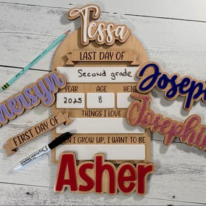 May include: Wooden school-themed signs and name cutouts. Includes a sign for "Last Day of Second Grade" with fill-in sections, plus names like Tessa, Joseph, and Asher. A pencil and marker are also present.