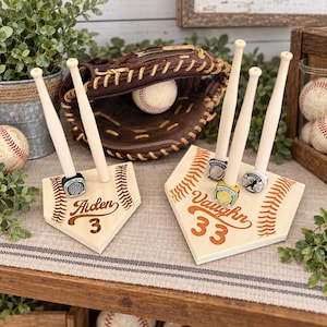 May include: Two light-colored wooden home plate displays featuring engraved baseball stitching, names Aiden and Vaughn, and jersey numbers 3 and 33. Each display holds miniature wooden baseball bats and championship rings.