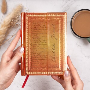Puede incluir: Un diario de gratitud de color dorado y naranja con un borde decorativo y las palabras "Gratitude Journal" escritas en la portada.