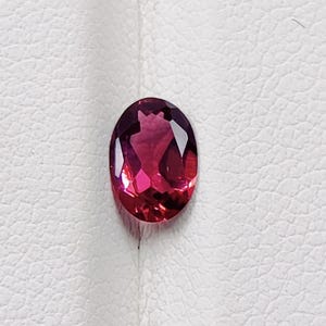 May include: An oval-shaped, faceted gemstone with a deep red and pink color gradient. The gem is set against a white, textured background, showcasing its polished surface and internal reflections. This gemstone is suitable for jewelry making.