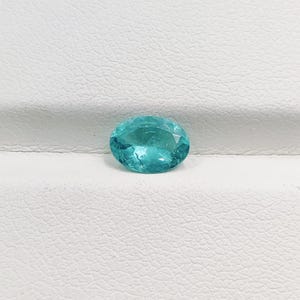 Pode incluir: Uma gema facetada, de forma oval, na cor verde-azulado. A gema tem um corte brilhante com m&uacute;ltiplas facetas que refletem a luz. A pedra &eacute; apresentada sobre um fundo branco, real&ccedil;ando sua cor e clareza. Adequada para joias.