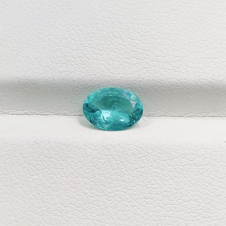 Pode incluir: Uma gema facetada, de forma oval, na cor verde-azulado. A gema tem um corte brilhante com m&uacute;ltiplas facetas que refletem a luz. A pedra &eacute; apresentada sobre um fundo branco, real&ccedil;ando sua cor e clareza. Adequada para joias.