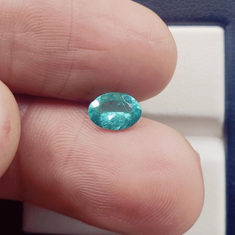 Pode incluir: Gema ovalada, cor verde-azulado, segurada entre os dedos. A gema tem uma superf&iacute;cie facetada, refletindo a luz. A cor &eacute; um verde-azulado m&eacute;dio e vibrante, com uma ligeira translucidez. A gema est&aacute; sobre um fundo neutro.