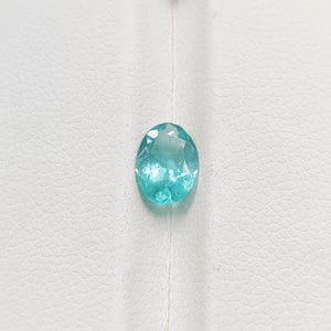 Pode incluir: Uma pedra preciosa ovalada azul clara com m&uacute;ltiplas facetas. A pedra preciosa est&aacute; colocada sobre um fundo branco texturizado. A gema tem uma apar&ecirc;ncia brilhante e transl&uacute;cida.