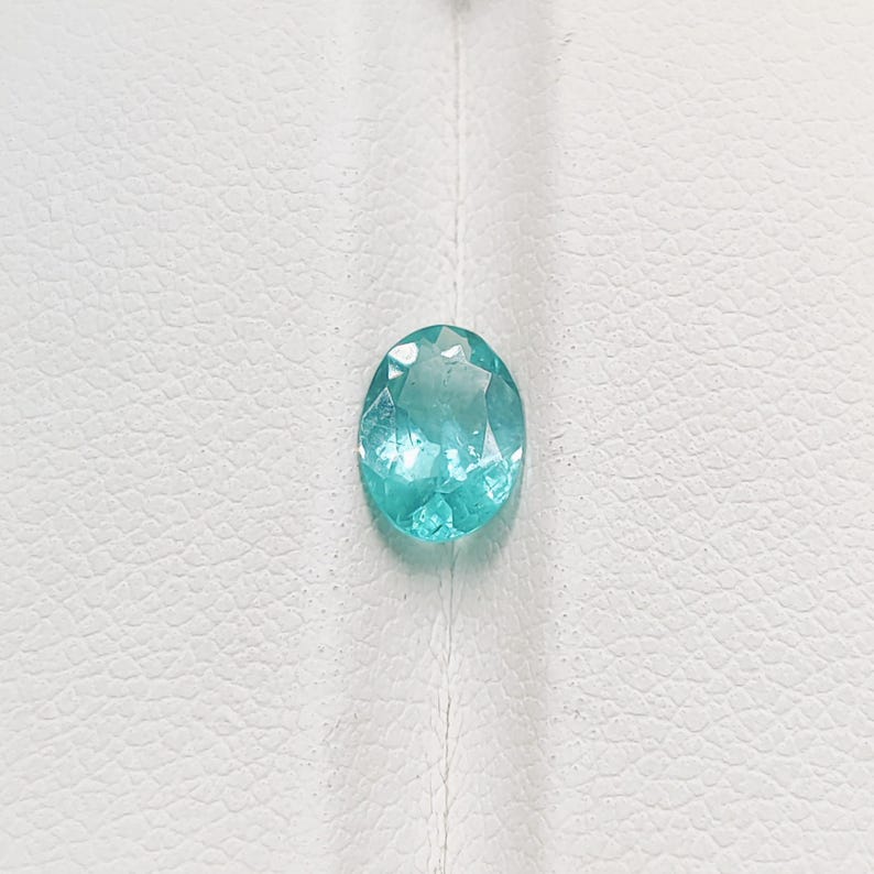 Pode incluir: Uma pedra preciosa ovalada azul clara com m&uacute;ltiplas facetas. A pedra preciosa est&aacute; colocada sobre um fundo branco texturizado. A gema tem uma apar&ecirc;ncia brilhante e transl&uacute;cida.