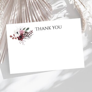 May include: A white card with the text "THANK YOU" printed in black. The card is decorated with a floral arrangement of white, pink, and burgundy flowers.