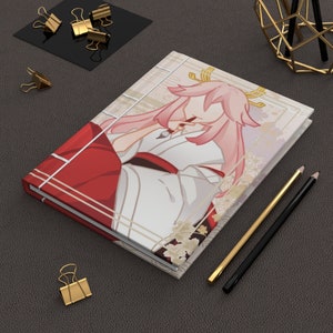 May include: A pink and white illustrated notebook with a character wearing a red and white kimono. The notebook has a gold and white patterned design.