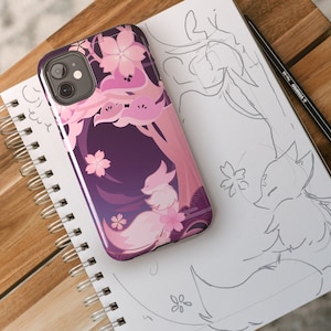 May include: A purple phone case with a pink and white fox design. The fox is surrounded by pink cherry blossoms. The case is on a white notebook with a pencil.
