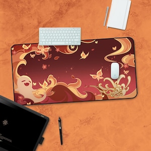 May include: A red and gold mousepad with a whimsical design featuring stylized clouds, butterflies, and a ghost-like figure. The mousepad is on a light orange surface with a white keyboard and a white notepad.
