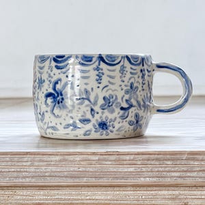 Puede incluir: Una taza de cerámica con base blanca y un estampado floral azul. La taza tiene forma redondeada con asa y está decorada con flores y hojas pintadas a mano. El diseño incluye un borde festoneado en la parte superior.