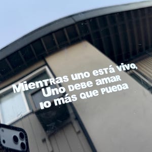 May include: White vinyl decal with the Spanish text "Mientras uno está vivo, Uno debe amar lo más que pueda" on a car window. The decal is in a sans-serif font. A phone case with a leopard print is reflected in the window.