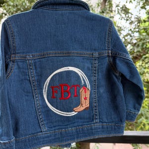May include: A blue denim jacket with a white rope circle design on the back. Inside the circle are the red letters "FBT" and a brown cowboy boot with a red star. The jacket has a button closure and two front pockets.