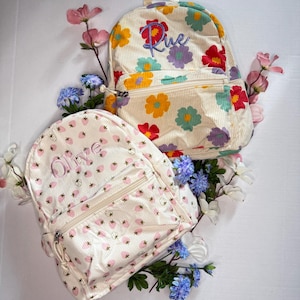 May include: Two white canvas backpacks with floral patterns. One backpack has a pink strawberry pattern and says "Olive" on the front. The other backpack has a multi-colored floral pattern and says "Rue" on the front.