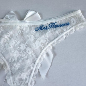 May include: White lace underwear with a floral pattern and satin bows. The words "Mrs. Herrera" are embroidered in blue script across the front. The edges are trimmed with a delicate scalloped lace.