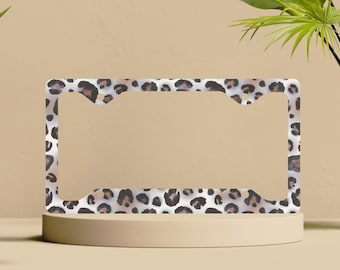 May include: License plate frame with a leopard print pattern. The frame features a white background with brown and black spots. The frame is rectangular with cutouts for the license plate and mounting hardware.