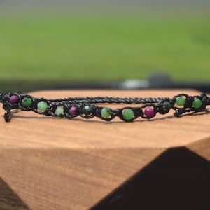 May include: A black cord bracelet featuring a series of small, faceted, round beads in alternating shades of vibrant green and deep magenta. The beads are secured within a knotted black string design that forms a continuous, flexible band.