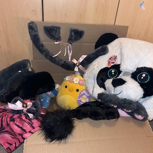 Könnte beinhalten: Eine Sammlung von Plüschtieren und Accessoires, darunter ein Pandabär, ein gelbes Küken mit Blumenkranz und ein rosa-schwarzes Outfit. Ebenfalls enthalten sind schwarze Katzenohren mit rosa Schleifen und ein flauschiger schwarzer Schwanz.