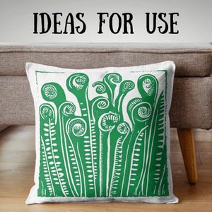 May include: White square cushion featuring a green fern design. The fern illustration is a detailed linocut style print. The words "IDEAS FOR USE" are printed in black above the cushion. The cushion is on a neutral-coloured sofa.