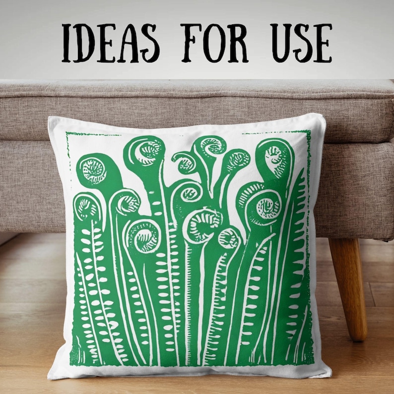 May include: White square cushion featuring a green fern design. The fern illustration is a detailed linocut style print. The words "IDEAS FOR USE" are printed in black above the cushion. The cushion is on a neutral-coloured sofa.