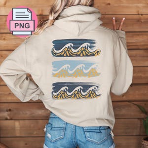 Puede incluir: Una sudadera con capucha beige con un gráfico de tres olas azules, amarillas y blancas en la espalda. Las olas están estilizadas con un efecto de pincelada.
