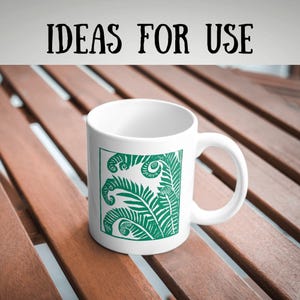 May include: White ceramic mug featuring a green fern design. The mug has a curved handle and the words "IDEAS FOR USE" in black text at the top. The mug is on a wooden surface.