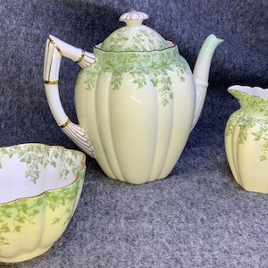 May include: A vintage tea set with a teapot, creamer, and sugar bowl. The set is cream-colored with green leaf patterns and gold accents. The teapot has a bamboo-style handle.