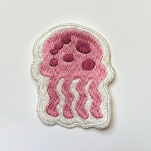 May include: Pink embroidered iron-on patch of a jellyfish with white outline. The jellyfish has pink spots and tentacles.