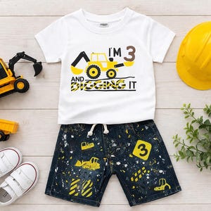 Könnte beinhalten: Ein weißes T-Shirt und Jeans-Shorts-Set für ein 3-jähriges Kind mit einem Baustellen-Thema. Das T-Shirt hat eine gelb-schwarze Bagger-Grafik mit dem Text "I'M 3 AND DIGGING IT". Die Shorts haben ein Farbspritzer-Design und Baufahrzeug-Grafiken.