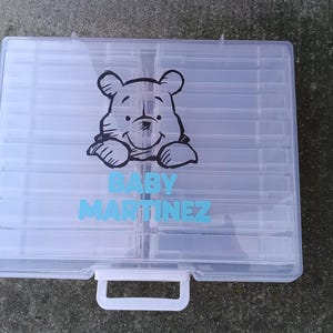 Könnte beinhalten: Eine transparente Kunststoff-Organizer-Box mit weißem Griff und einer Cartoon-Bär-Grafik. Der Text "BABY MARTINEZ" ist blau unter der Bärenillustration aufgedruckt. Die Box ist in mehrere Fächer unterteilt.