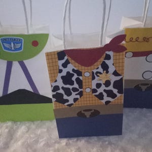 May include: Three themed gift bags. One is green and white with a Space Ranger logo, another is designed as a cowboy with a cow-print vest, and the third is yellow with a red design. All have white handles.