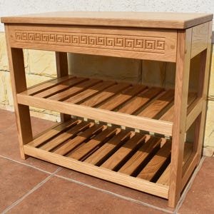 May include: A light brown wooden console table featuring a rectangular top and two slatted shelves below. The front apron of the table is decorated with a carved Greek key pattern. The piece is constructed with four vertical legs and horizontal supports.
