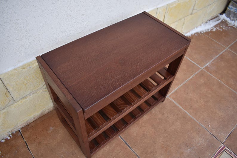 May include: A rectangular dark brown wooden shoe rack featuring a solid flat top surface and two lower slatted shelves for storage. The unit is constructed with sturdy vertical side panels and horizontal supports, showcasing a natural wood grain finish.