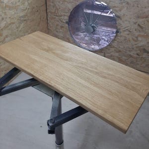 May include: A rectangular light brown wooden board with a visible natural grain pattern resting on a dark gray metal support stand. The board has smooth, flat surfaces and straight edges, positioned horizontally in a workshop environment.