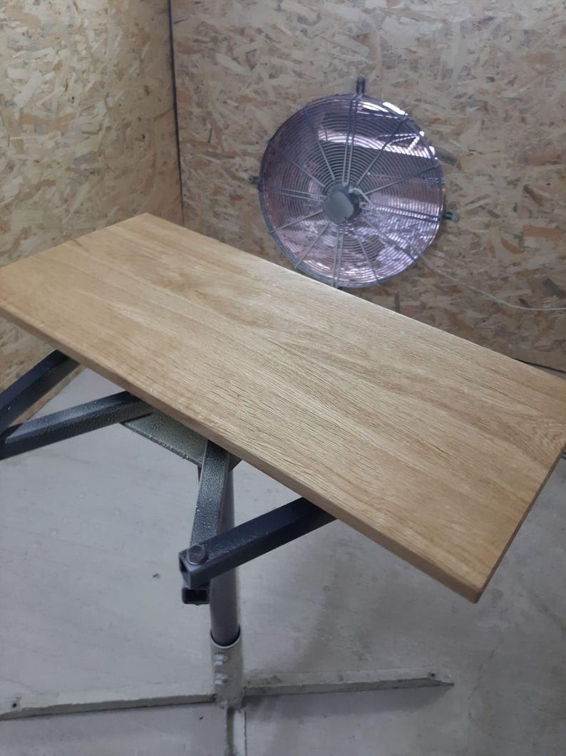 May include: A rectangular light brown wooden board with a visible natural grain pattern resting on a dark gray metal support stand. The board has smooth, flat surfaces and straight edges, positioned horizontally in a workshop environment.