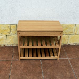 May include: A light brown wooden shoe rack featuring a solid rectangular top surface and two lower slatted shelves. The unit is constructed with four vertical legs and horizontal support beams, providing an open storage design for footwear.