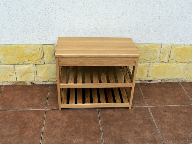 May include: A light brown wooden shoe rack featuring a solid rectangular top surface and two lower slatted shelves. The unit is constructed with four vertical legs and horizontal support beams, providing an open storage design for footwear.