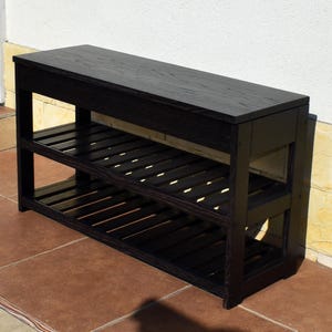 May include: A rectangular wooden bench finished in a dark black stain. The furniture piece features a solid flat top surface and two lower tiers constructed with horizontal slats, providing open storage space across the entire length of the unit.