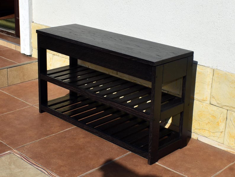 May include: A rectangular wooden bench finished in a dark black stain. The furniture piece features a solid flat top surface and two lower tiers constructed with horizontal slats, providing open storage space across the entire length of the unit.