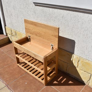 May include: A light brown wooden shoe rack with a rectangular storage compartment featuring a hinged lid that opens upward. The unit includes two slatted shelves below the storage area, providing open space for footwear organization and storage.