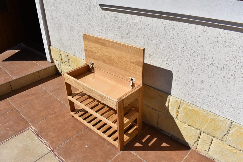 May include: A light brown wooden shoe rack with a rectangular storage compartment featuring a hinged lid that opens upward. The unit includes two slatted shelves below the storage area, providing open space for footwear organization and storage.