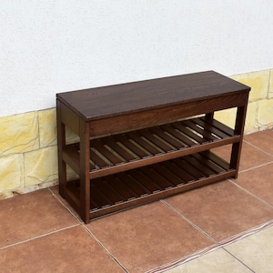 May include: A dark brown wooden shoe rack featuring a solid rectangular top surface and two lower slatted shelves. The furniture piece has a sturdy frame with four vertical legs and horizontal supports, providing an open storage design.