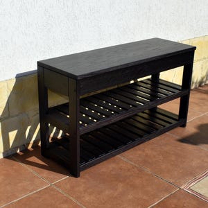 May include: A rectangular black wooden bench featuring a solid flat top surface and two lower slatted shelves. The furniture piece is constructed with four vertical legs and horizontal supports, showcasing a dark wood grain finish throughout the structure.
