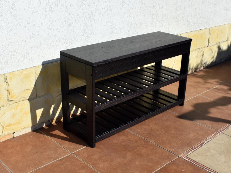 May include: A rectangular black wooden bench featuring a solid flat top surface and two lower slatted shelves. The furniture piece is constructed with four vertical legs and horizontal supports, showcasing a dark wood grain finish throughout the structure.